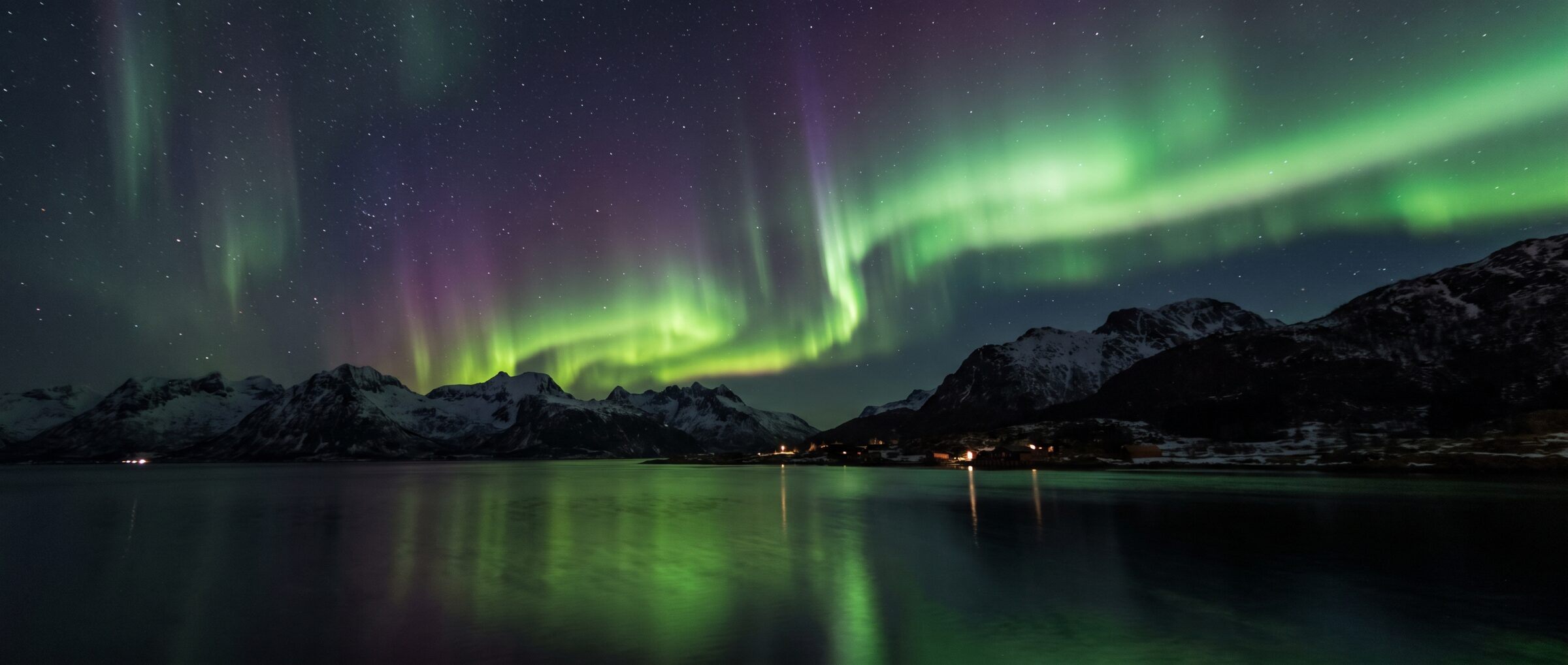 The aurora borealis in green and violet over a Norwegian fjord and snow-capped mountains.